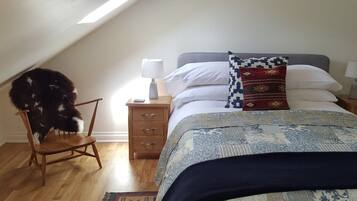 Et soveværelse med skrå vægge, en seng med mønstret sengetøj, en stol, et sengebord med en lampe og en lille måtte.