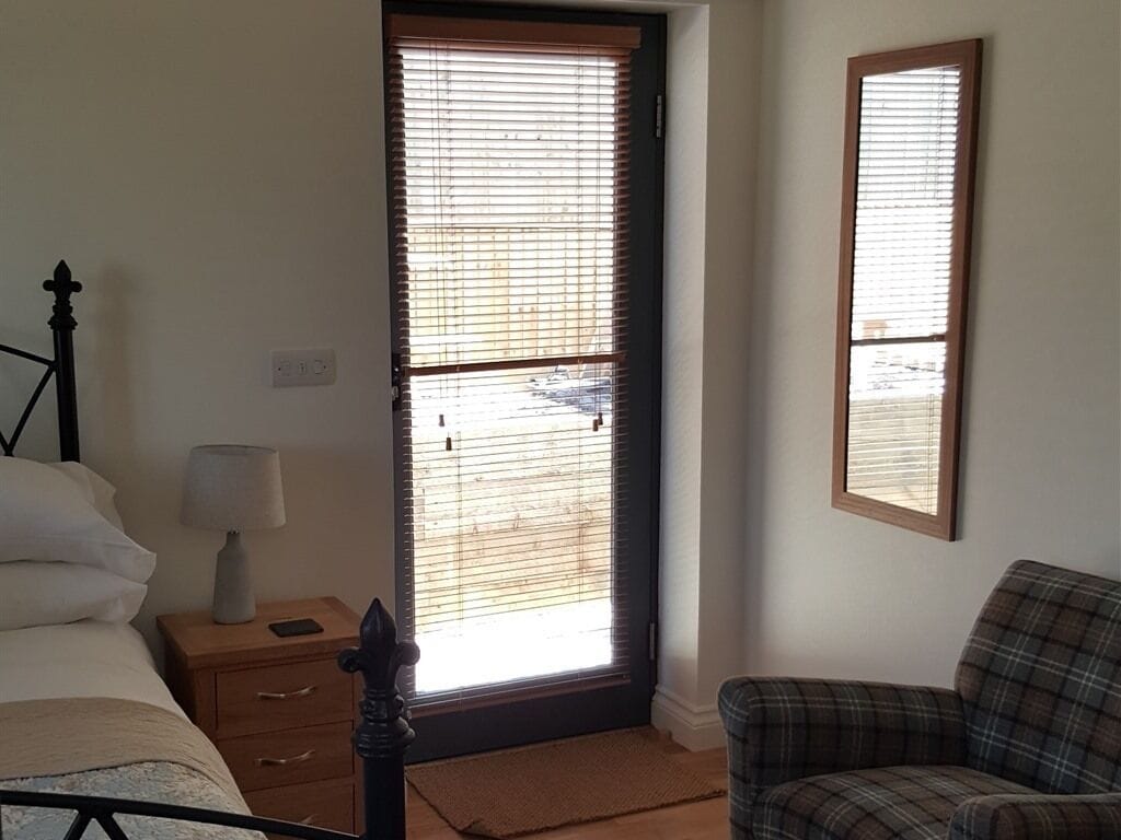 Une chambre à coucher avec un lit, une table de chevet avec une lampe, une chaise et une fenêtre avec des stores.
