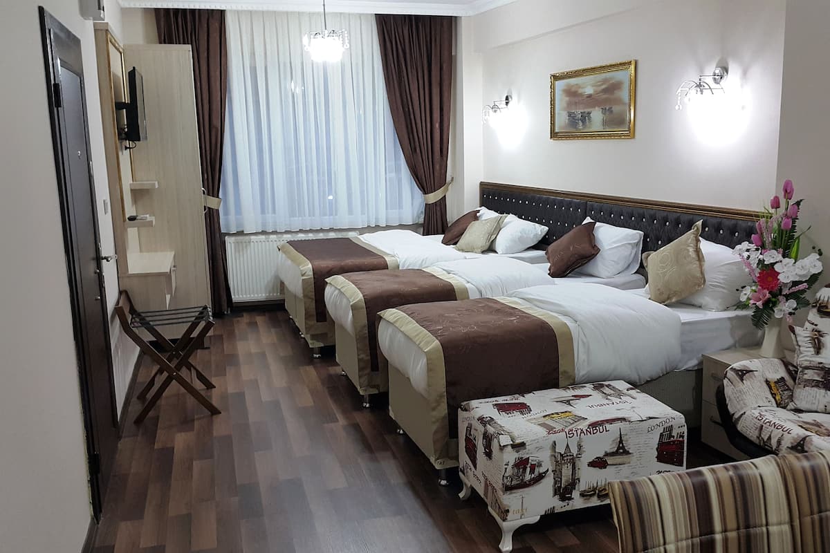 Une chambre d’hôtel avec trois lits, un coin salon avec un fauteuil, un radiateur, une fenêtre avec des rideaux et un tableau accroché au mur.