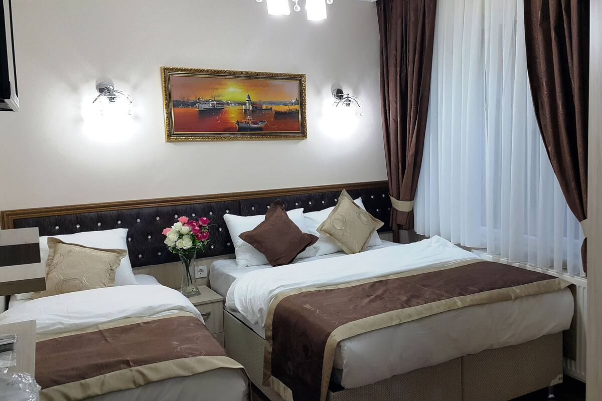 Une chambre d’hôtel avec deux lits, une table de chevet ornée d’un vase de fleurs, un tableau au mur et une fenêtre avec des rideaux.