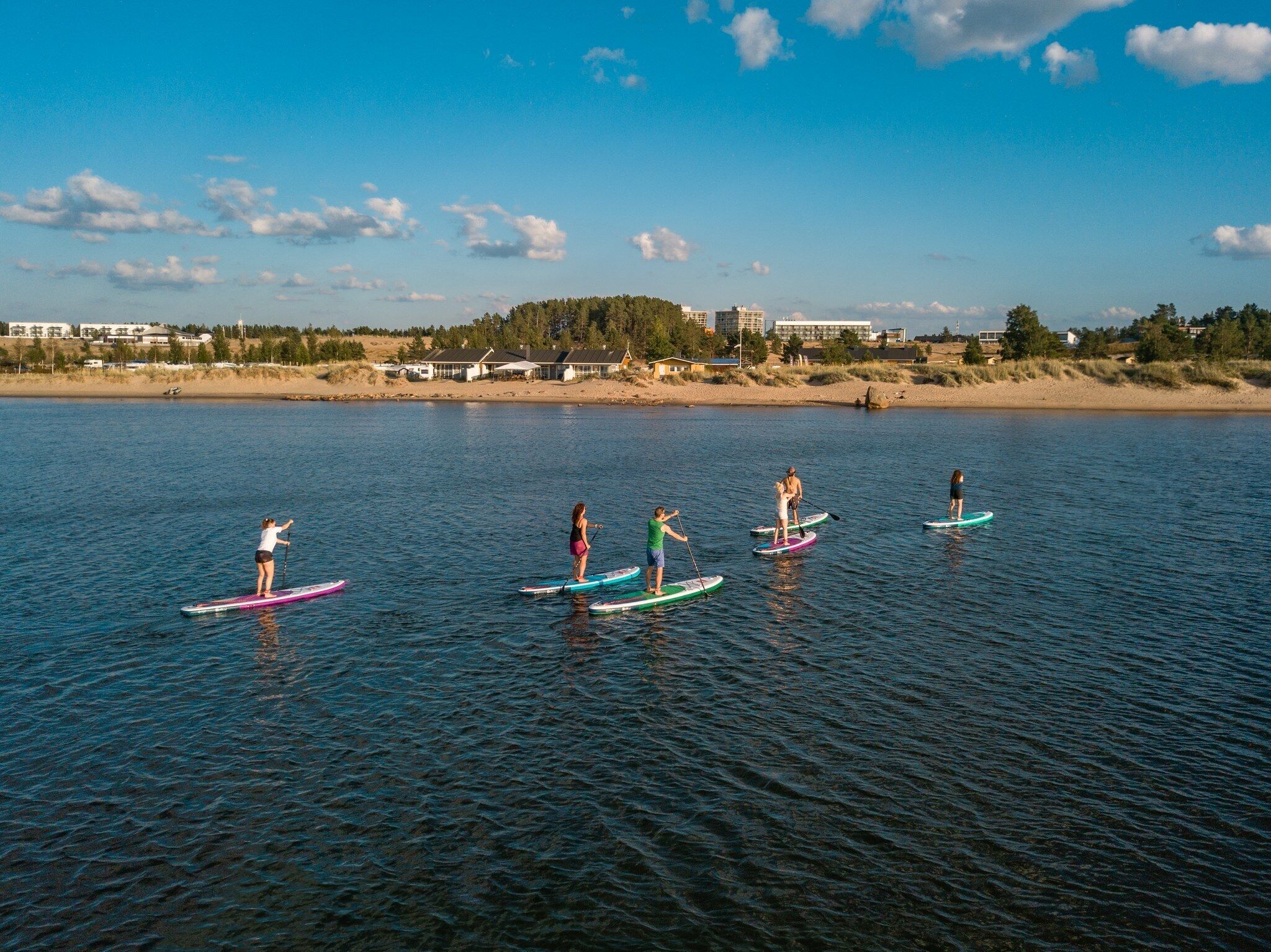 Eine Gruppe von Menschen steht auf Paddleboards auf ruhigem Wasser, mit einem Strand und Gebäuden im Hintergrund.