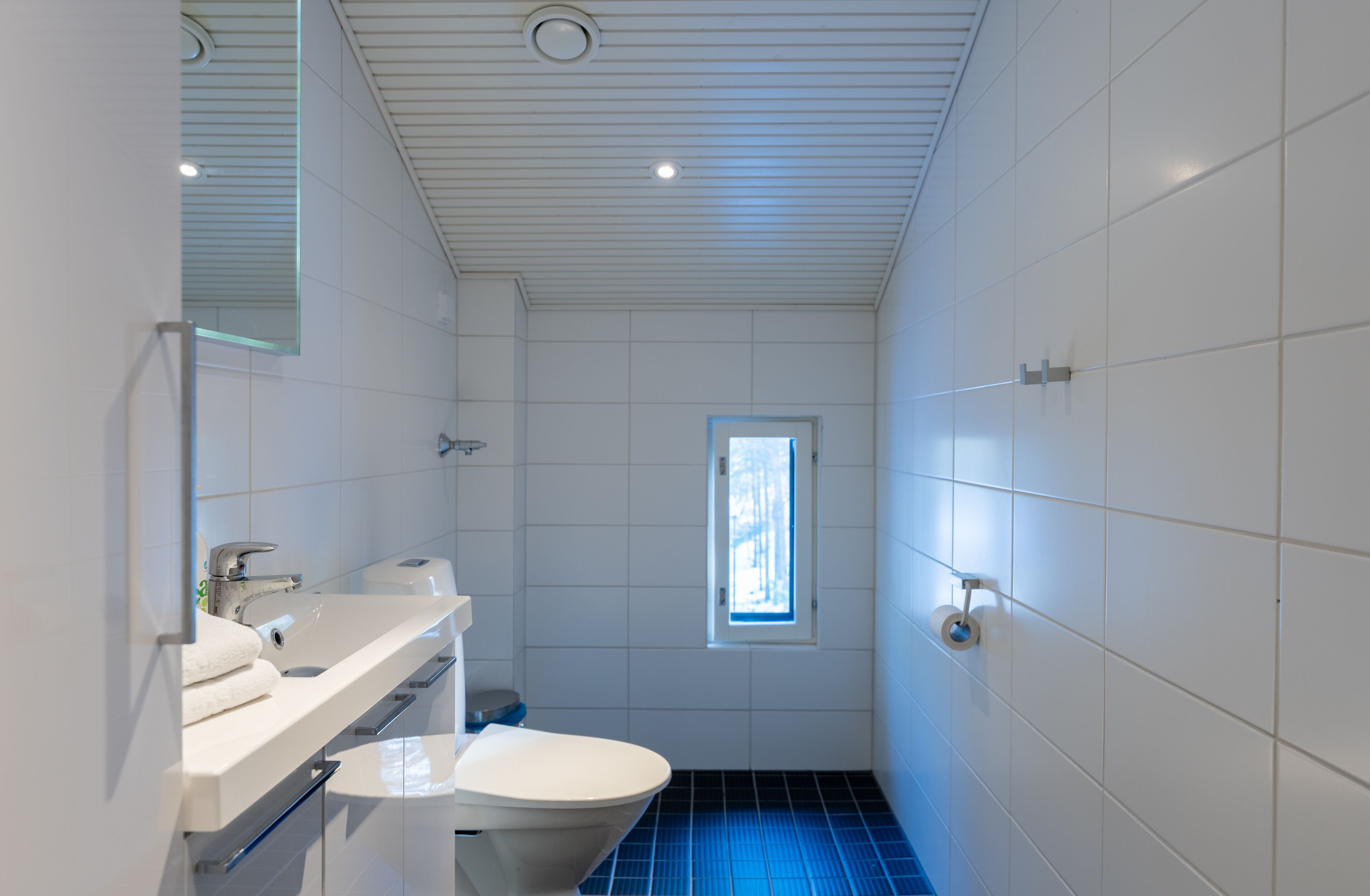 A modern bathroom with white tiles, a toilet, a sink, and a window.