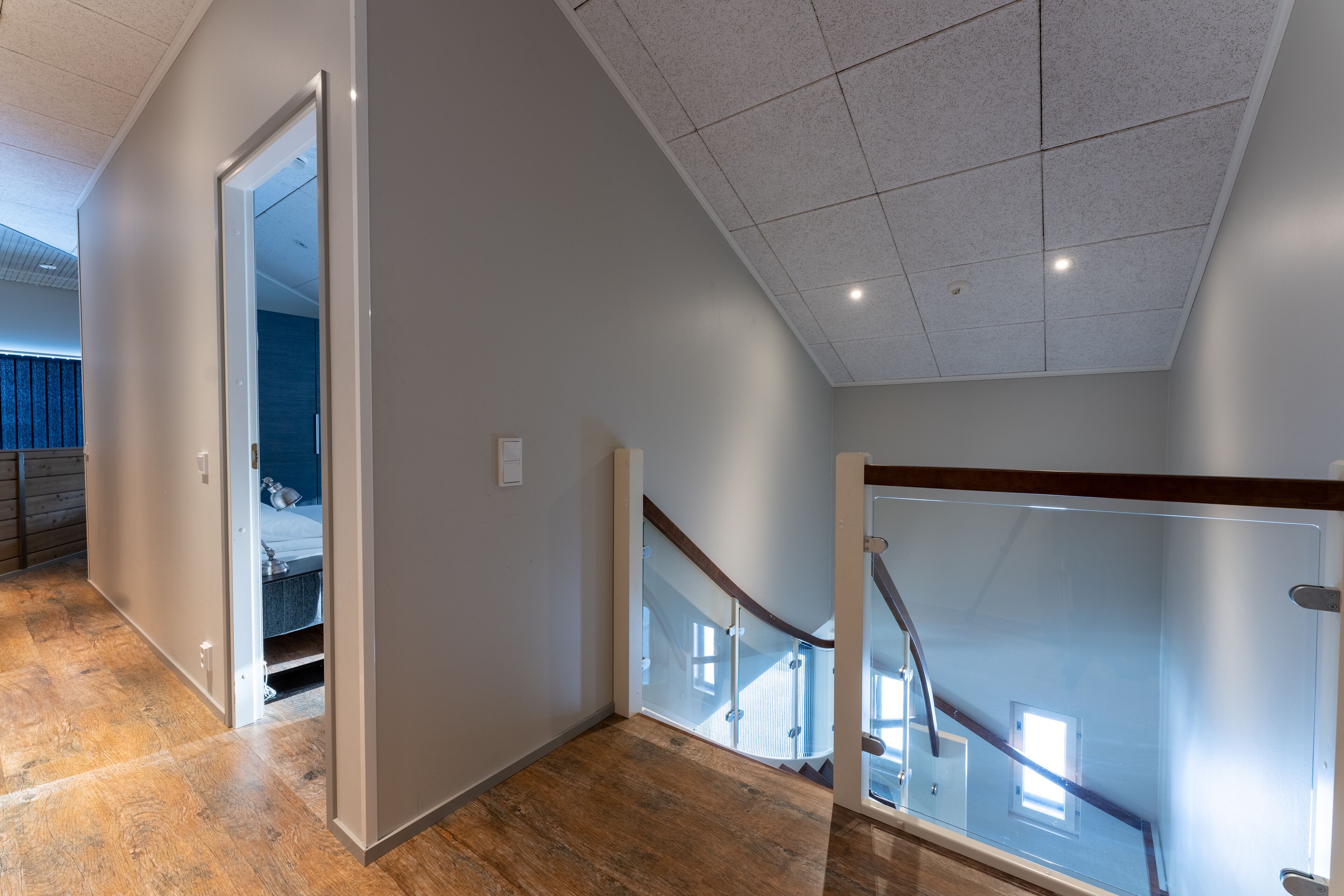 A spacious hallway with a wooden floor, a glass door leading to a bedroom, and a staircase with a glass railing.