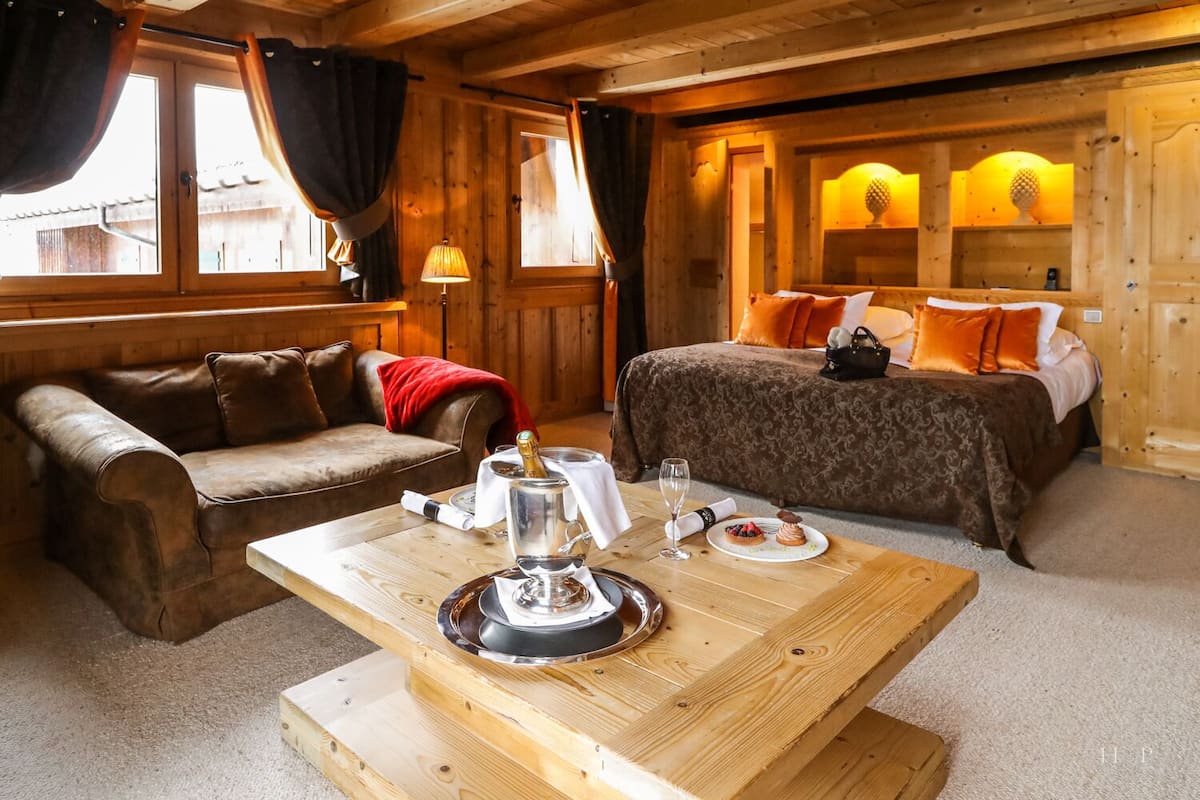 A cozy room with a wooden interior, a bed with orange pillows, a wooden table with a bottle and glasses, and a leather sofa.