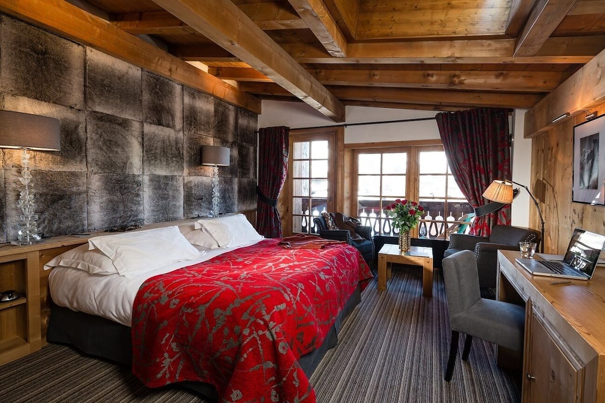 A bedroom with a large bed, a desk, and a chair. There is a window with curtains and a view of the outside.