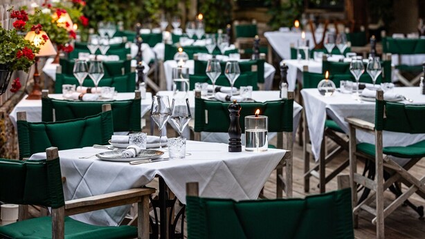 Un restaurante con mesas al aire libre, sillas verdes, manteles blancos y velas.