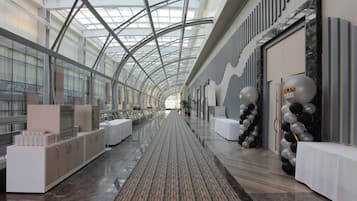 Ein geräumiger Korridor mit hoher Decke und Glasverkleidungen, der einen zentralen Gehweg und auf beiden Seiten weiße Tische zeigt.