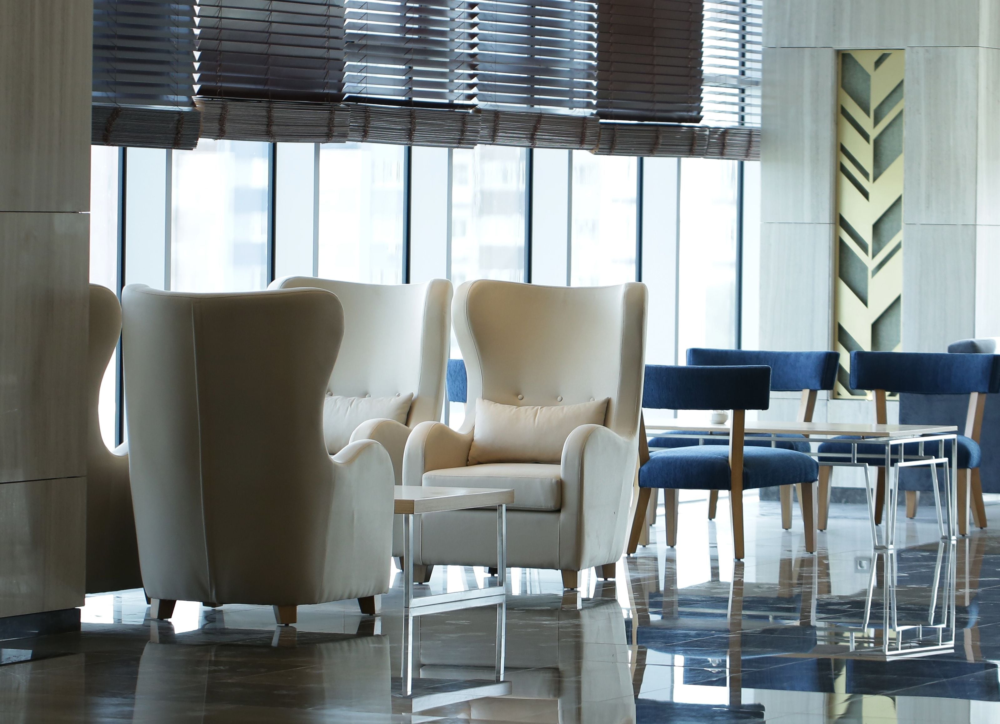 A modern lobby with large windows, a seating area with beige and blue chairs, and a reflective floor.