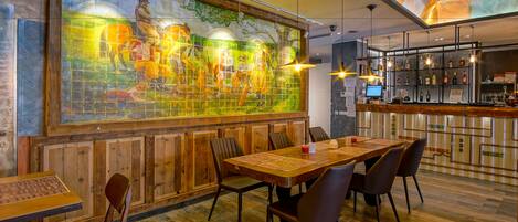 A restaurant with a large mural, wooden tables and chairs, and a bar area with shelves of bottles.