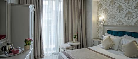 A hotel room with a bed, bedside table, lamp, curtains, and a desk with a kettle and teapot.