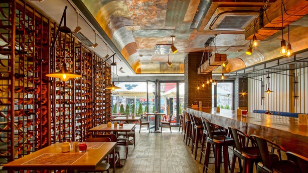 Un interior de restaurante con mesas y sillas de madera, una zona de bar y vistas a la calle a través de grandes ventanales.