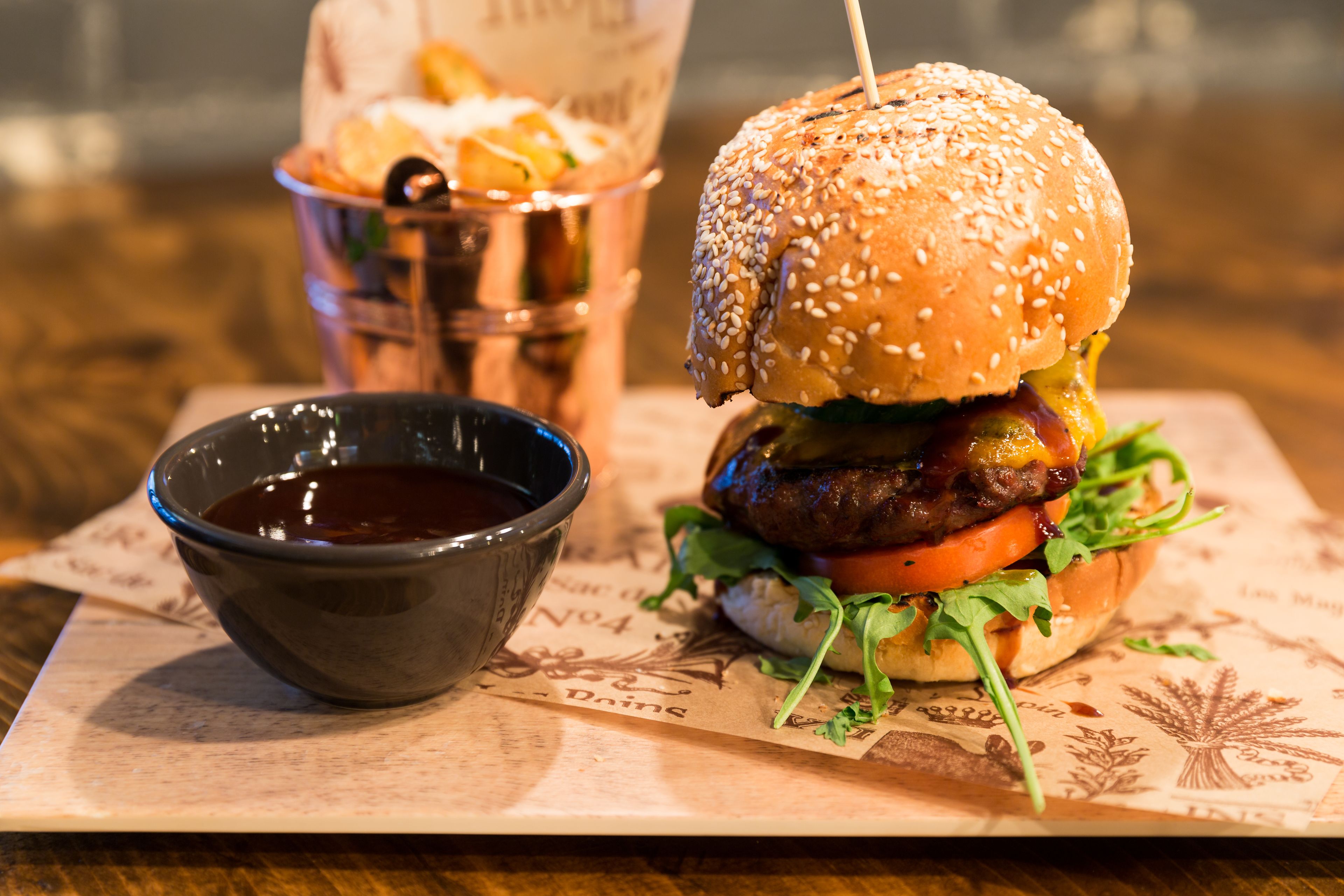 A burger with a sesame seed bun, lettuce, tomato, and a side of fries in a metal container, served on a paper plate with a small bowl of sauce.