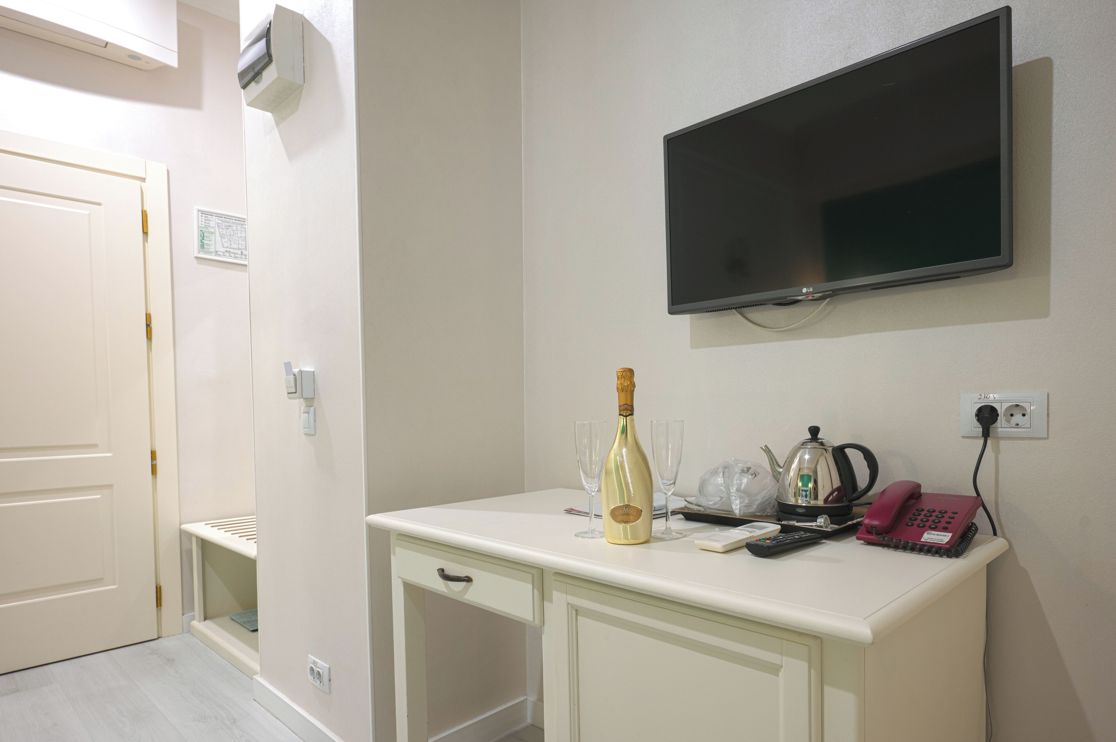A hotel room with a white desk, a bottle of champagne, two glasses, a telephone, and a television mounted on the wall.
