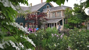 A two-story house with a porch, surrounded by a garden with various plants and flowers. - Holman's Heritage Suites (Summerside)