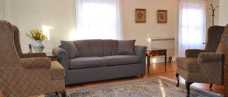 Un salon avec un canapé gris, deux fauteuils, un tapis à motifs et un radiateur.
