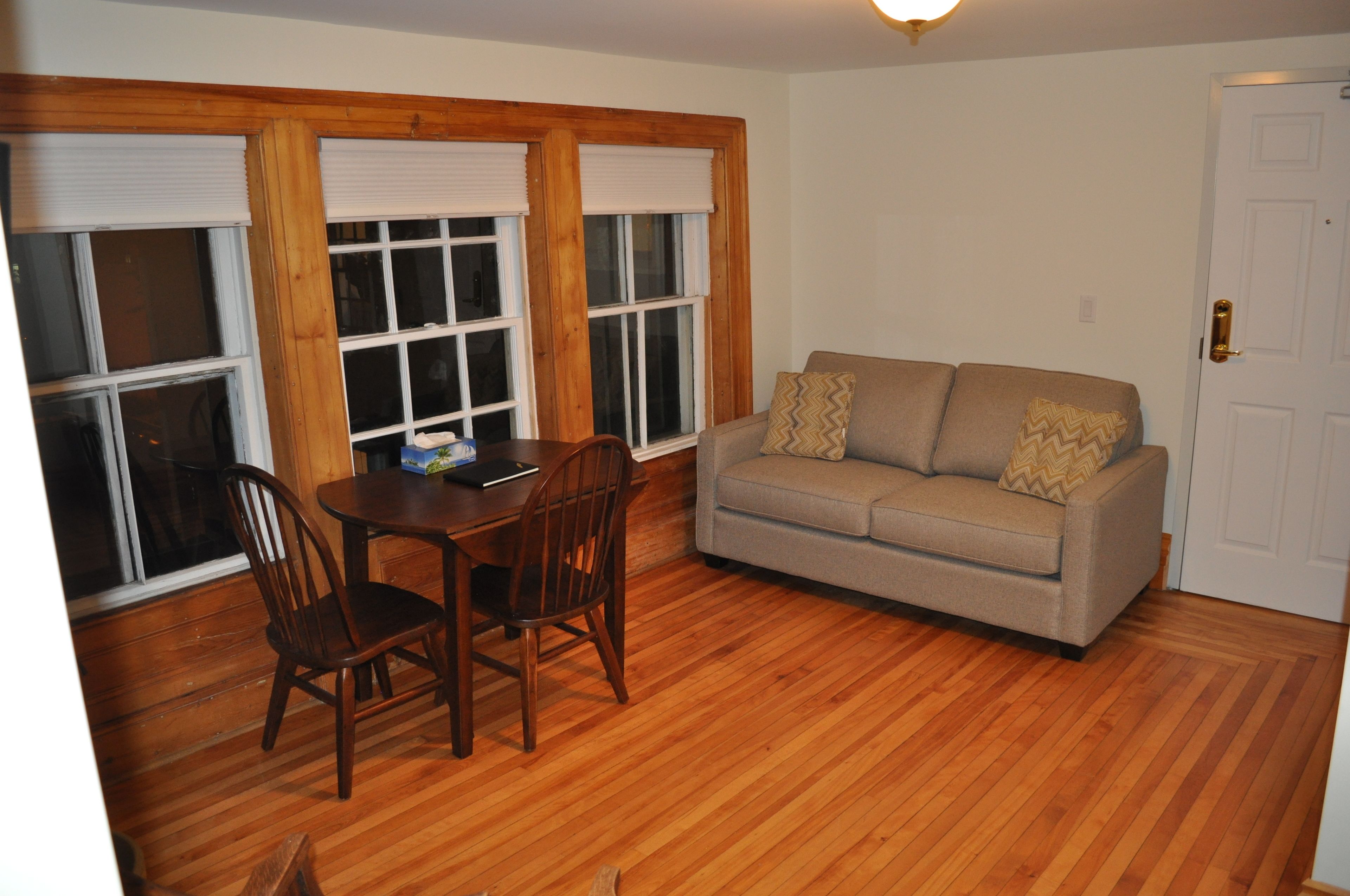 A room with a wooden floor, a beige sofa with patterned cushions, a dining table with chairs, and large windows with blinds.