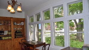 A dining area with a wooden table and chairs, a bar with shelves of bottles, and large windows offering a view of a garden and street. - Holman's Heritage Suites (Summerside)