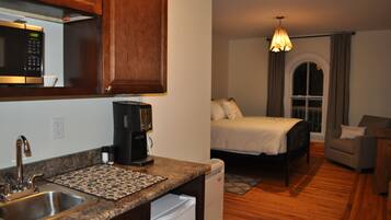 A compact hotel room with a kitchenette, a bed, a sofa, and a window with curtains.
