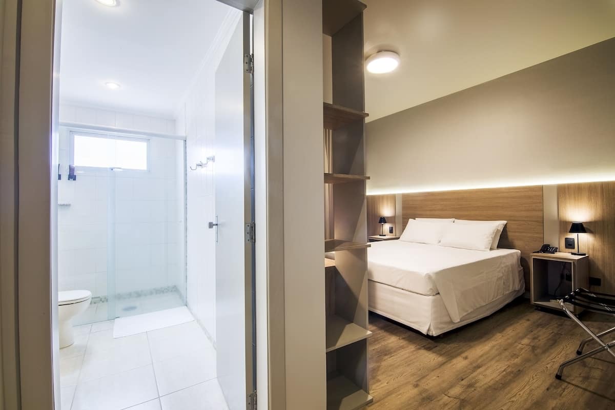 A hotel room with a shower, a bed, a bedside table with a lamp, and a wooden floor.