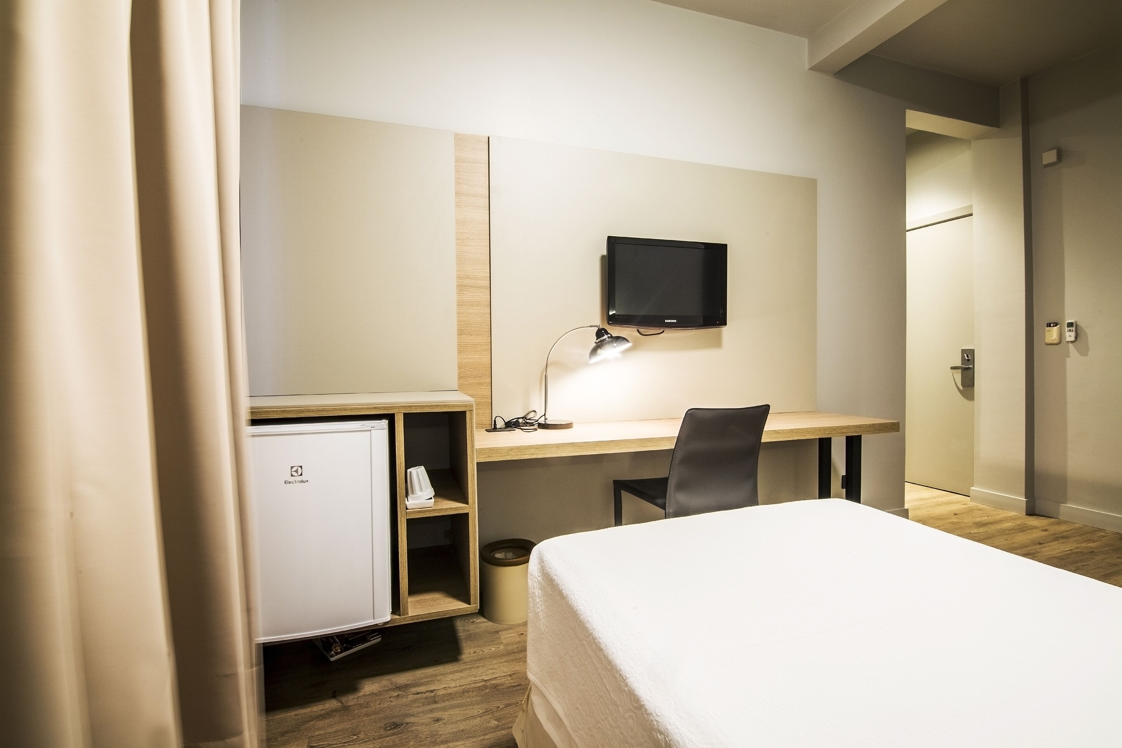 A hotel room with a bed, a desk with a computer and lamp, a TV mounted on the wall, and a small refrigerator.