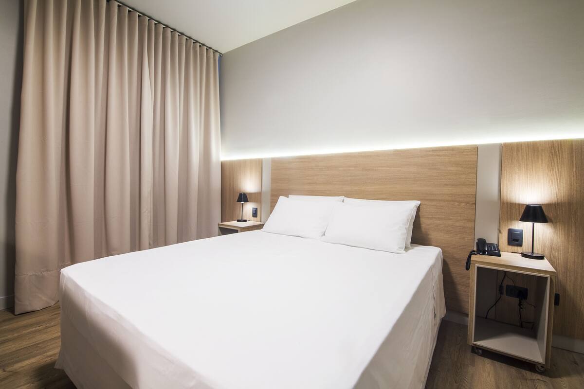 A hotel room with a large bed, two bedside tables with lamps, a wooden headboard, and a curtain on the window.
