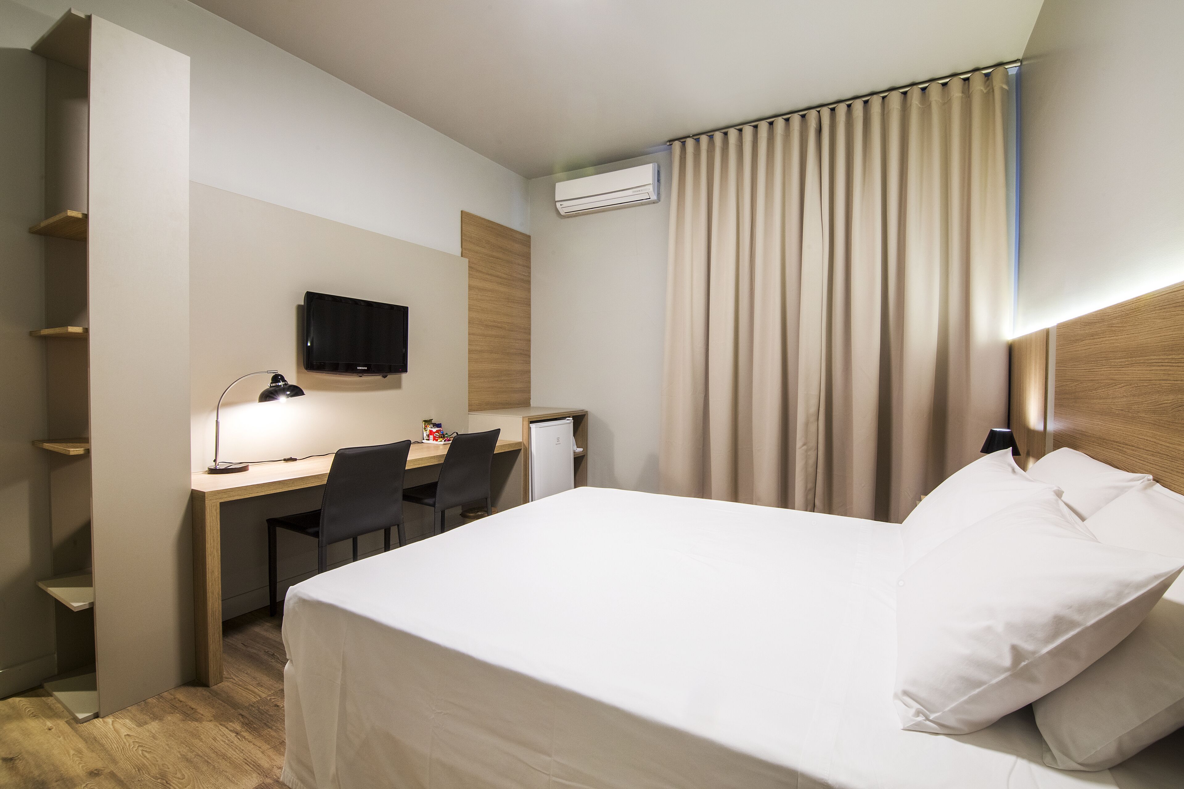 Una camera d'albergo con un letto, una scrivania con una sedia, una televisione e un condizionatore d'aria.