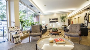 Una sala de estar moderna con una mesa de centro de vidrio, dos sillones, un sofá y un televisor. Se ven plantas, una alfombra y un atril para revistas.