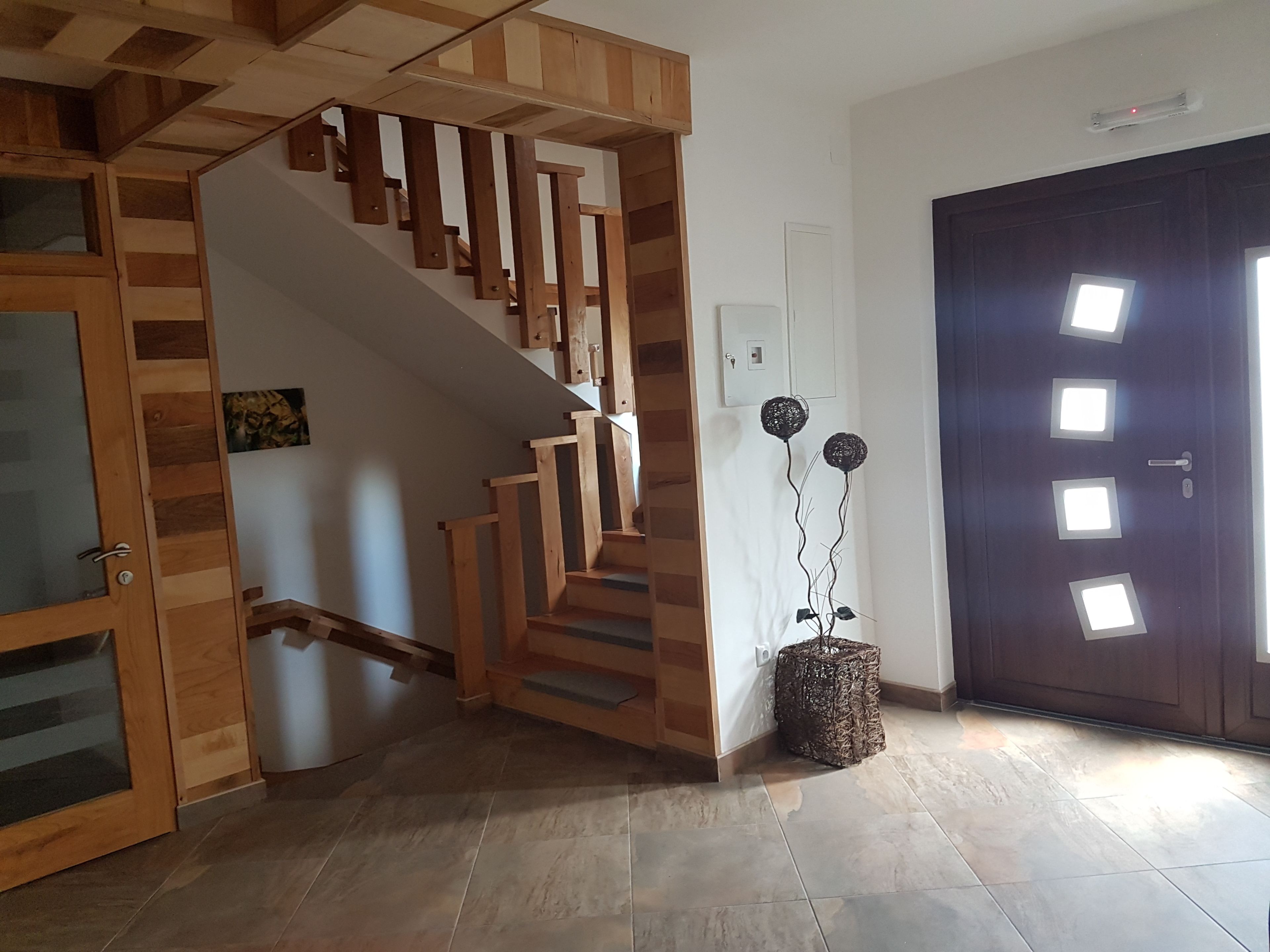 Una escalera de madera con una puerta de vidrio a la izquierda, una puerta oscura con recortes cuadrados a la derecha y una pequeña planta decorativa en el suelo.