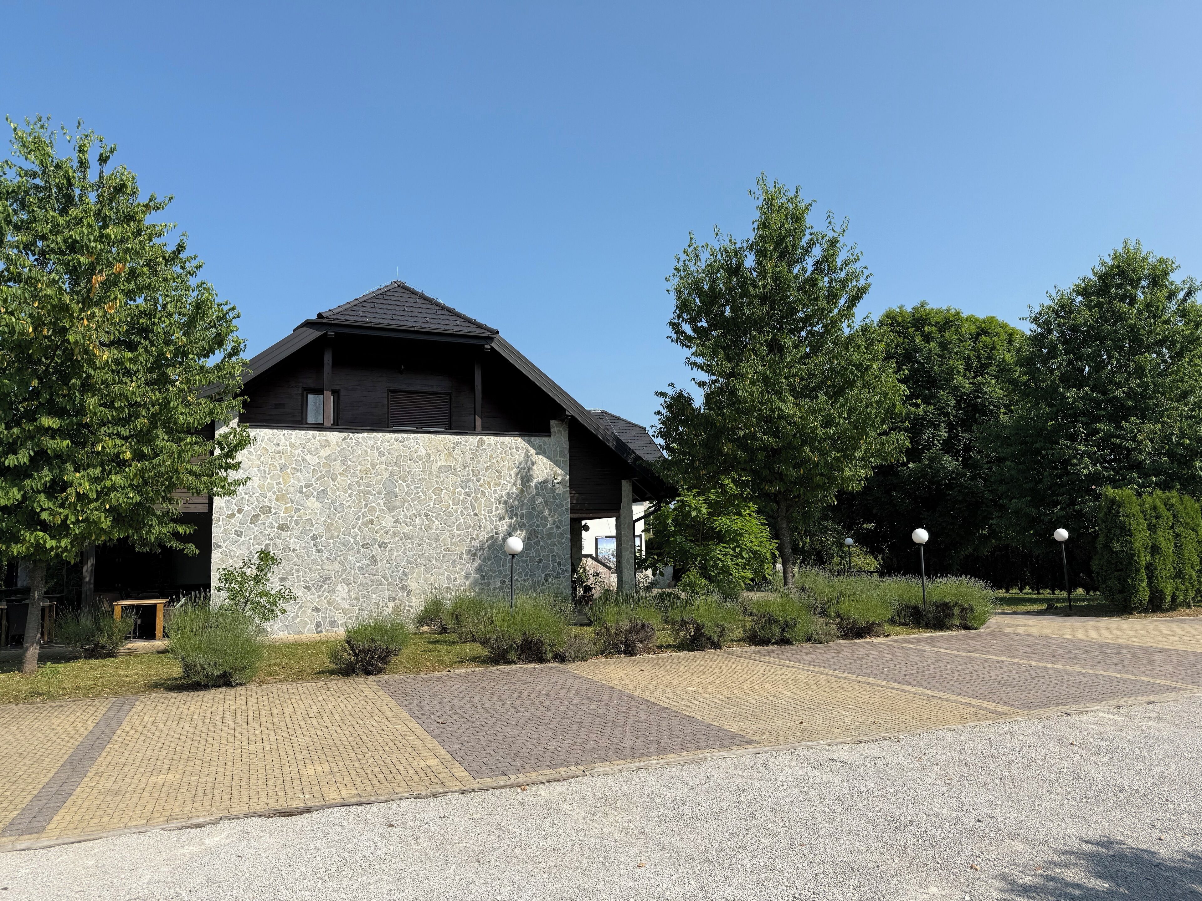 A building with a stone facade and a dark roof, surrounded by greenery and a paved area.