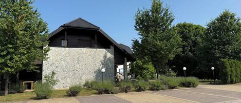 A building with a stone facade and a dark roof, surrounded by greenery and a paved area.
