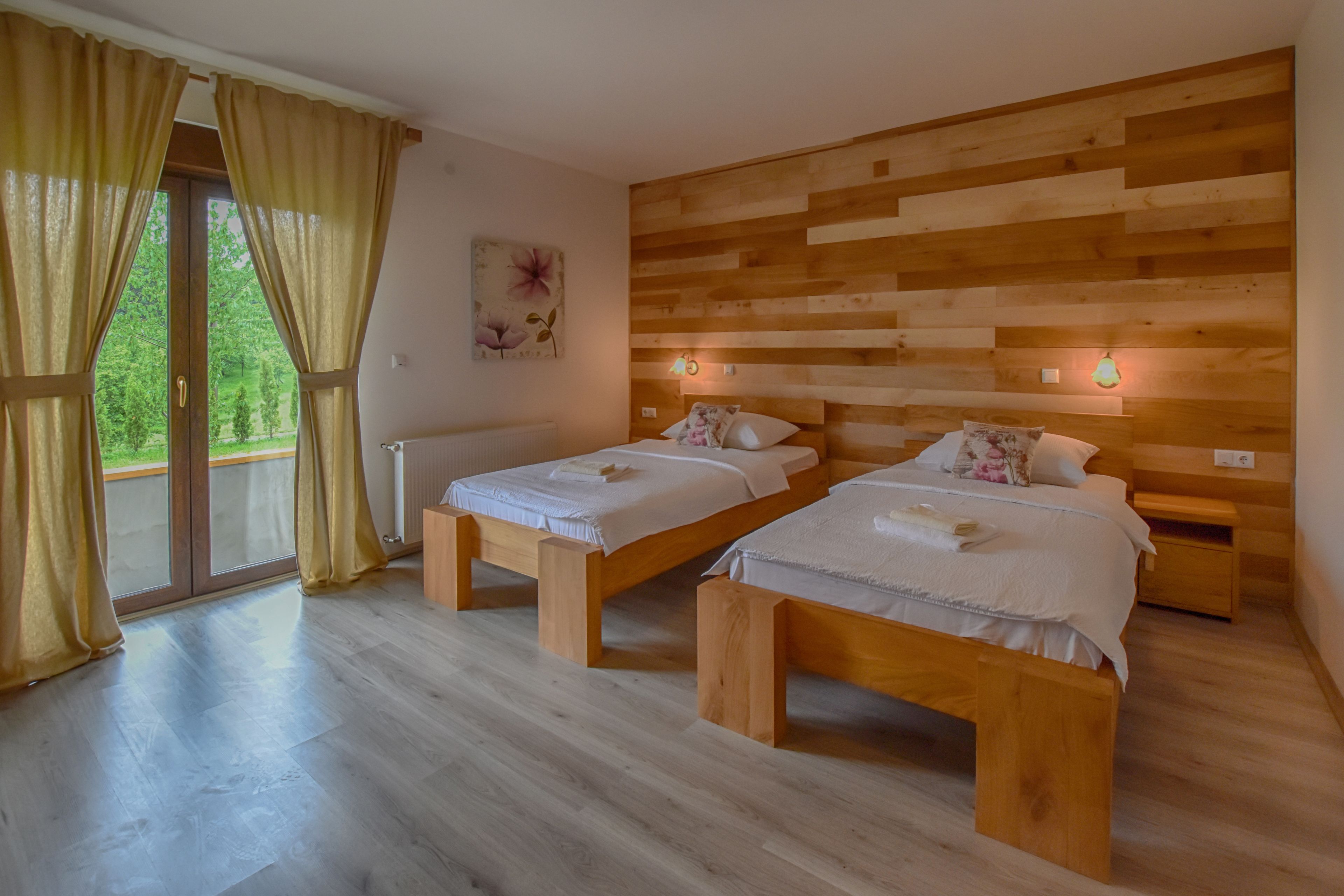 A room with two wooden beds, a wooden headboard, a large window with curtains, and a view of greenery outside.