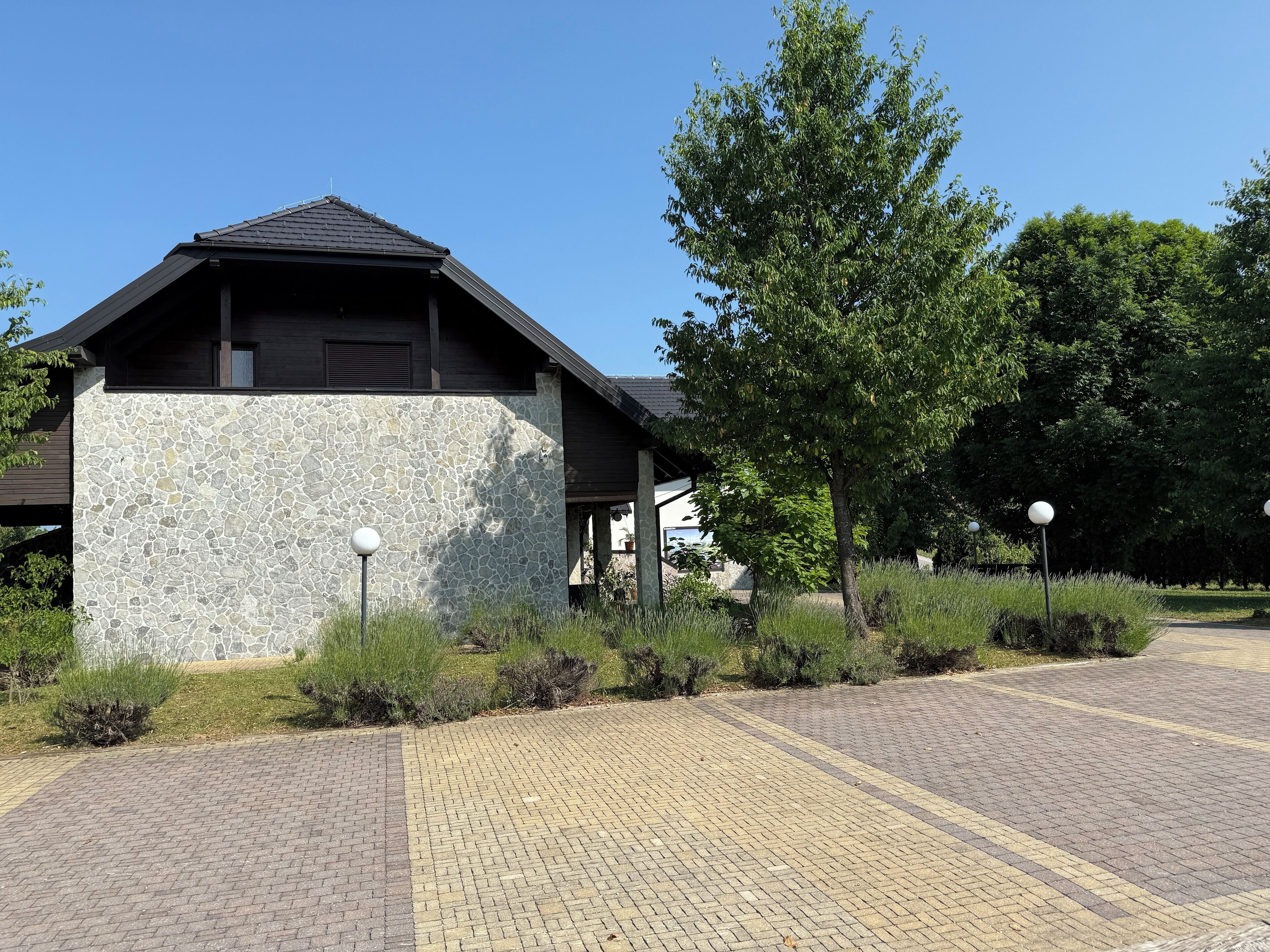 A building with a stone facade and a sloped roof, surrounded by greenery and a paved walkway.