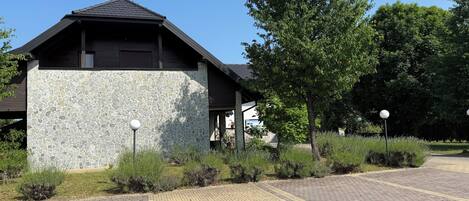 A building with a stone facade and a sloped roof, surrounded by greenery and a paved walkway.