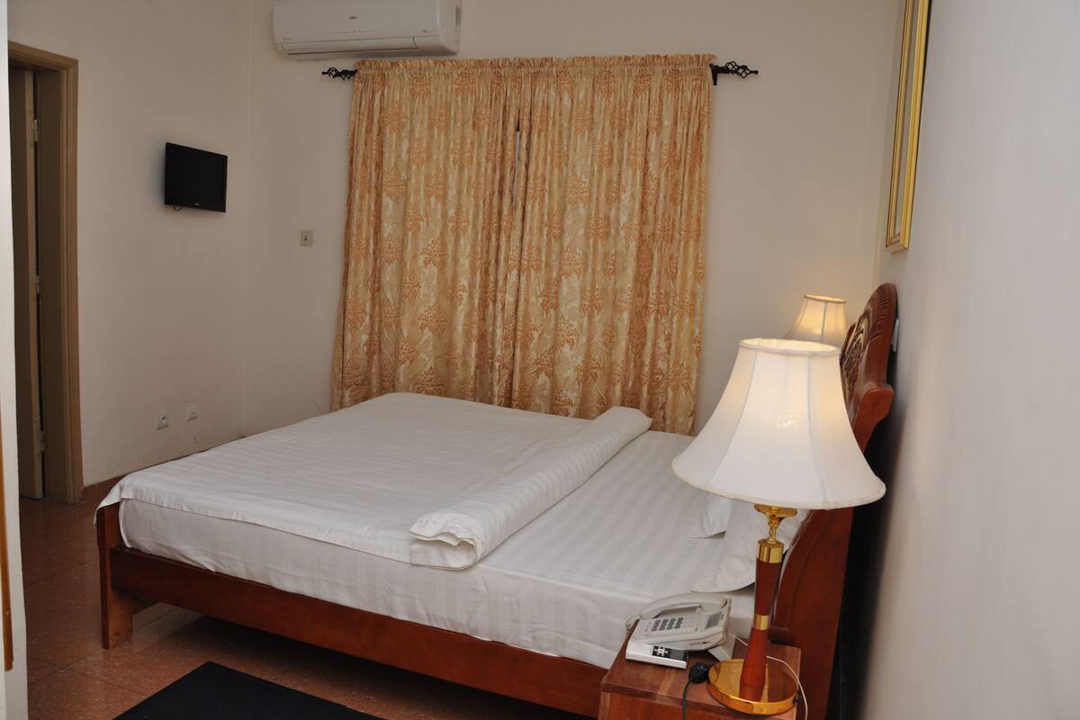 Cama bem arrumada com edredom branco e toalha dobrada, abajur, telefone e ar-condicionado fixado na parede.