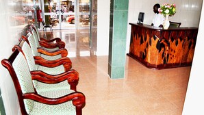 A reception area with a wooden reception desk, a chair with a green and white patterned cushion, and a vase with flowers. - Hotel Magnificat (Lomé)