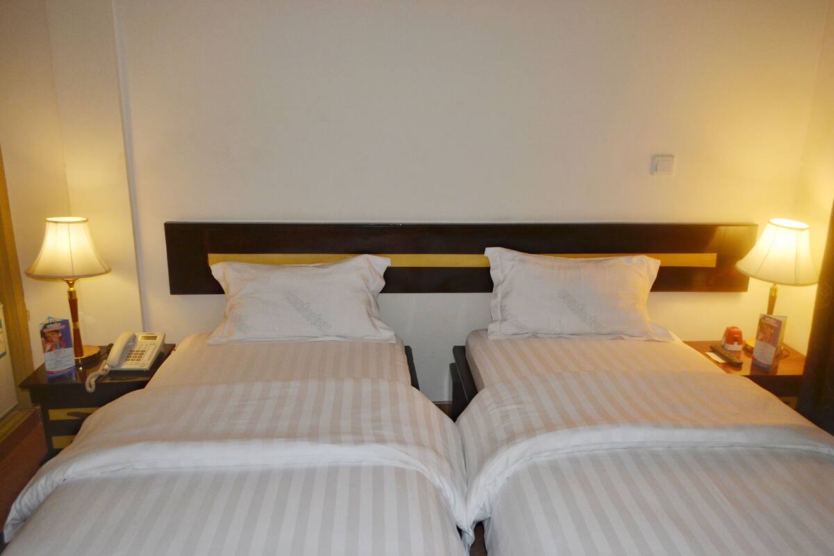 Quarto de hotel com duas camas, cada uma com roupa de cama branca e cabeceira de madeira. Há luminárias e telefone nas mesas de cabeceira.