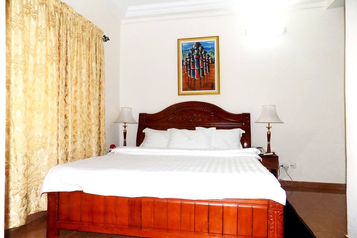 Quarto de hotel com cama de madeira, roupa de cama branca, um quadro na parede e duas luminárias de cabeceira.