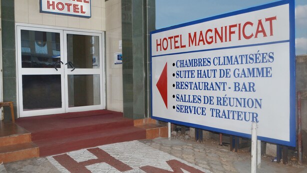 A hotel entrance with a sign indicating amenities such as a gym, high room, bar, meeting rooms, and a concierge service. - Hotel Magnificat (Lomé)