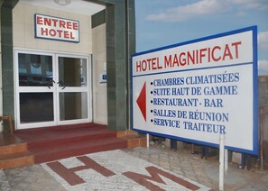 L’entrée d’un hôtel, avec une enseigne indiquant les équipements et services proposés, tels qu’une salle de sport, des chambres situées aux étages supérieurs, un bar, des salles de réunion et un service de conciergerie.