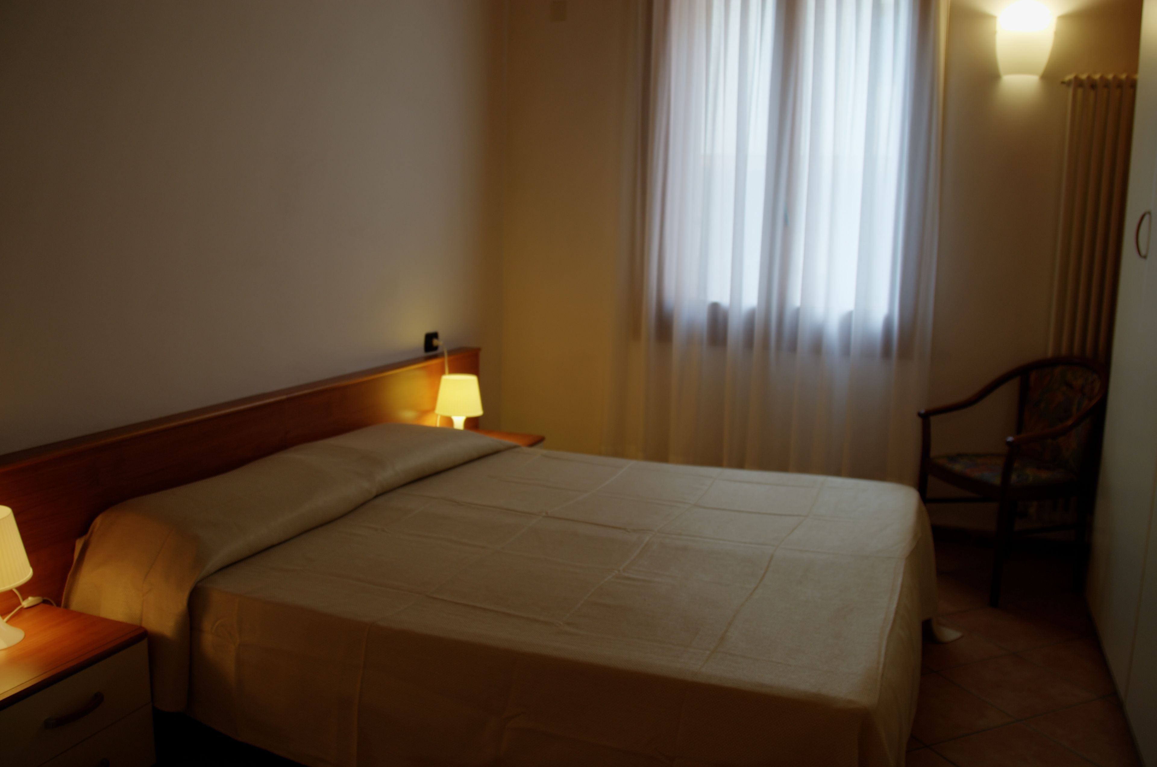 Hotellihuone, jossa on sänky, yöpöytä, lamppu, tuoli ja ikkuna, jossa on verhot.