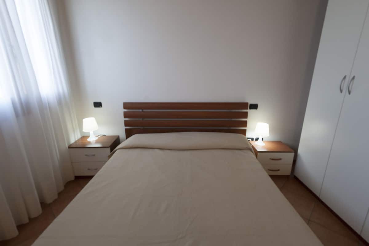A bedroom with a wooden headboard, two bedside tables with lamps, a bed with a beige bedspread, and a wardrobe.