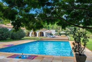 Una piscina con borde de baldosas, rodeada de vegetación y un edificio con arcos al fondo.