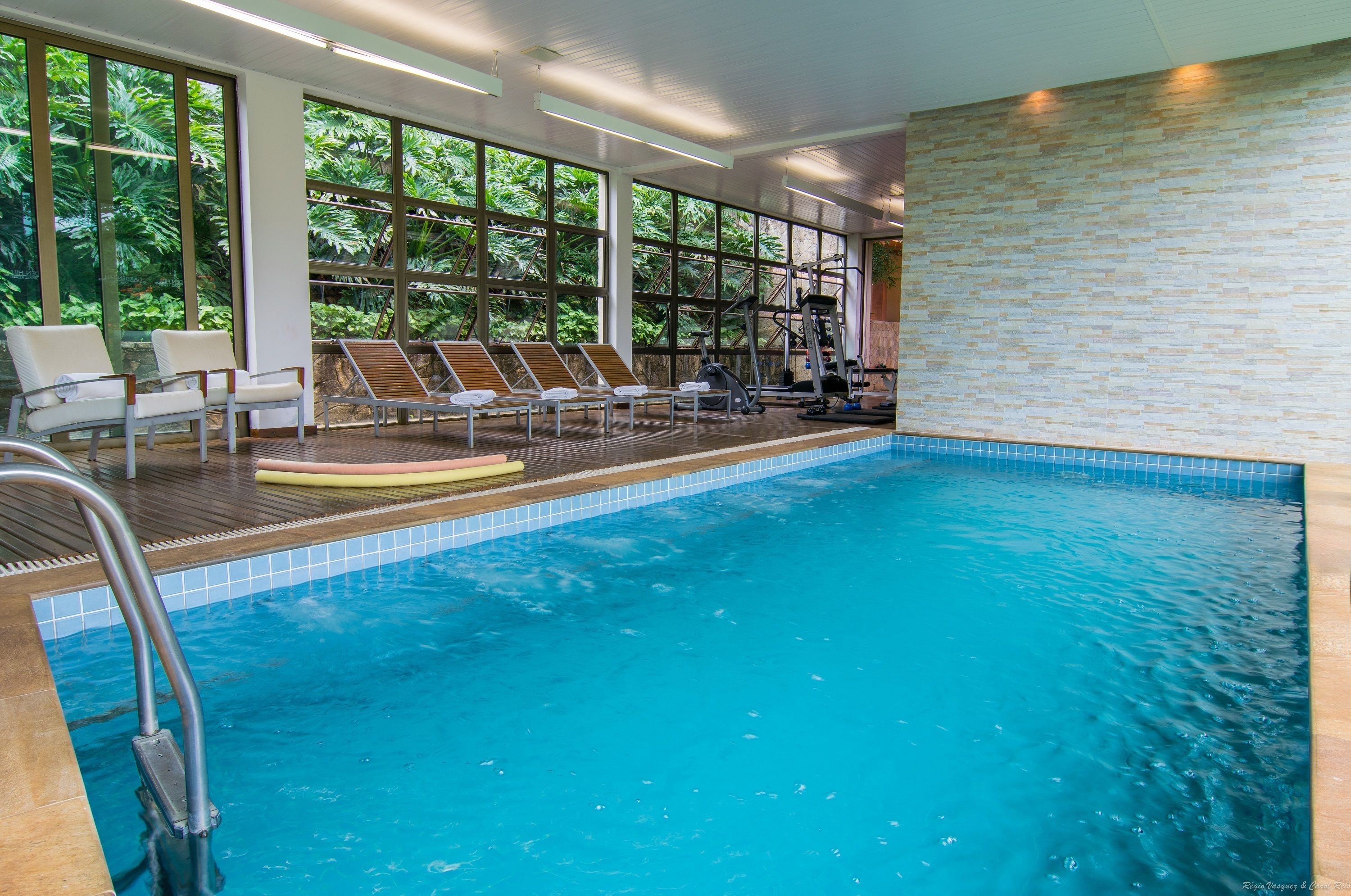 An indoor swimming pool with exercise equipment, sun loungers, and large windows offering a view of greenery.
