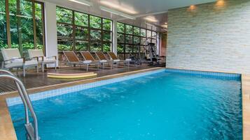 An indoor swimming pool with exercise equipment, sun loungers, and large windows offering a view of greenery.