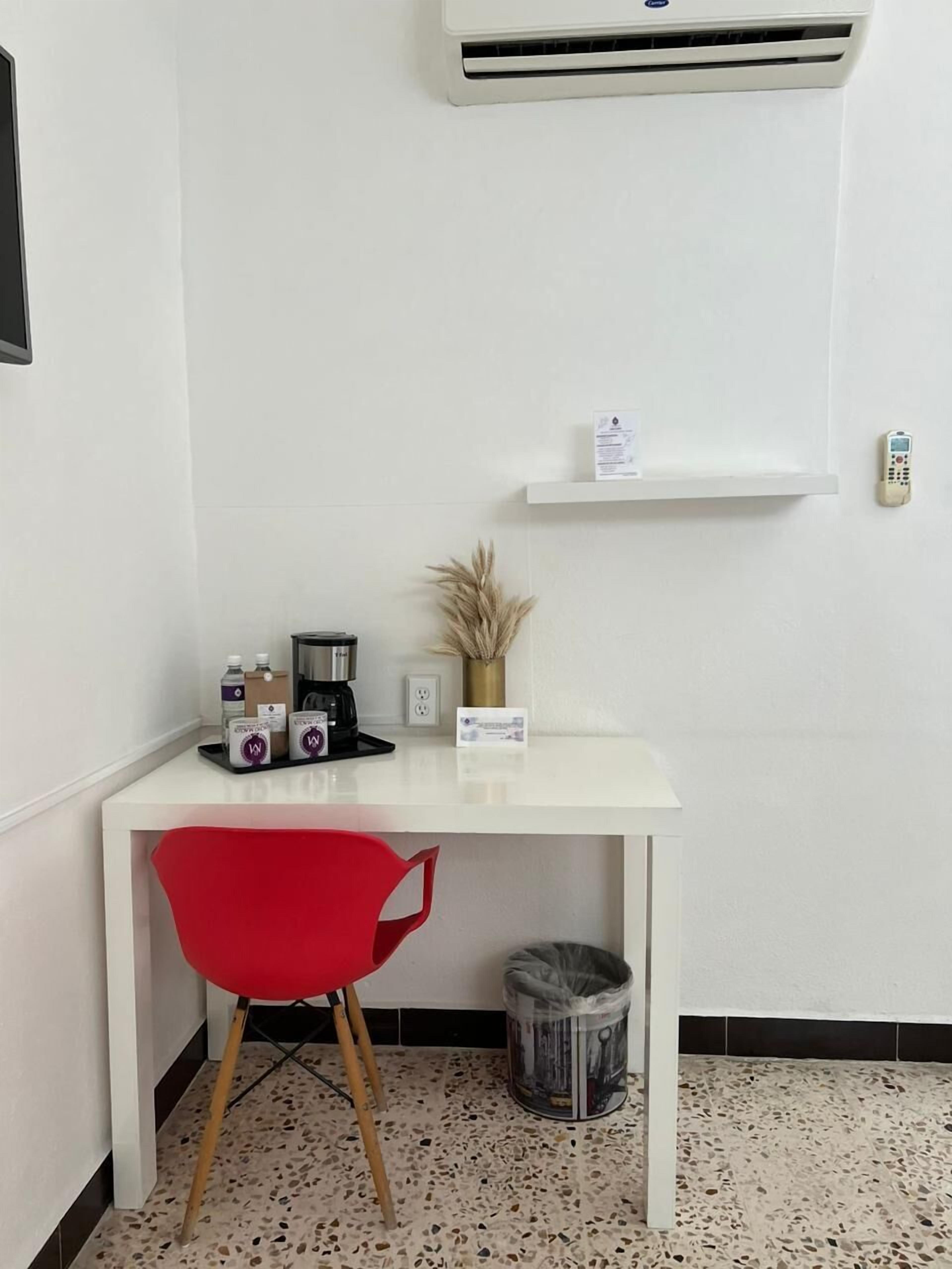 Un pequeño escritorio blanco con una silla roja, una cafetera, un florero con plantas secas y un cubo de basura.
