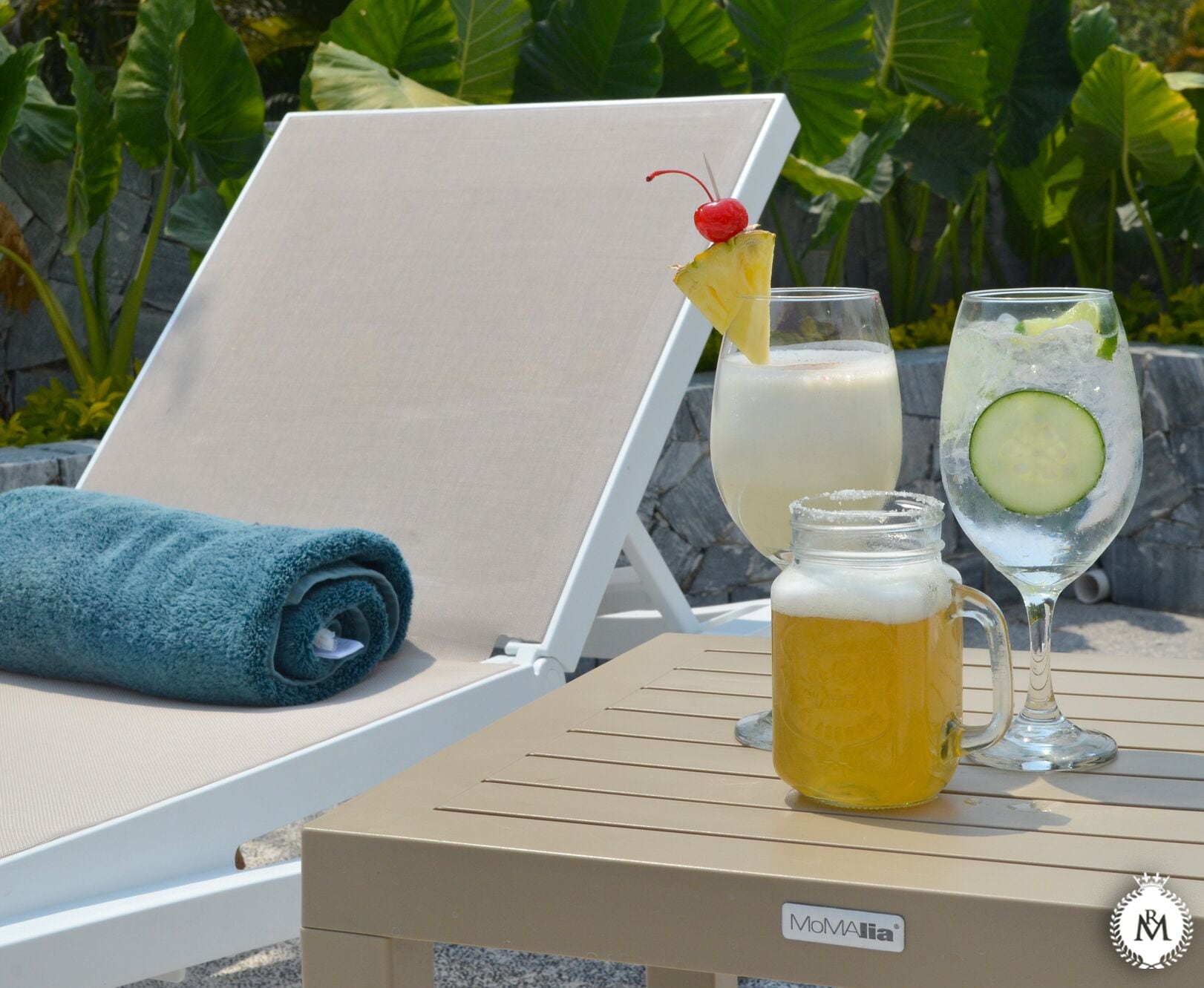 A sun lounger with a blue towel, a glass of iced drink with a pineapple slice and cherry, and a mug of beer on a table.