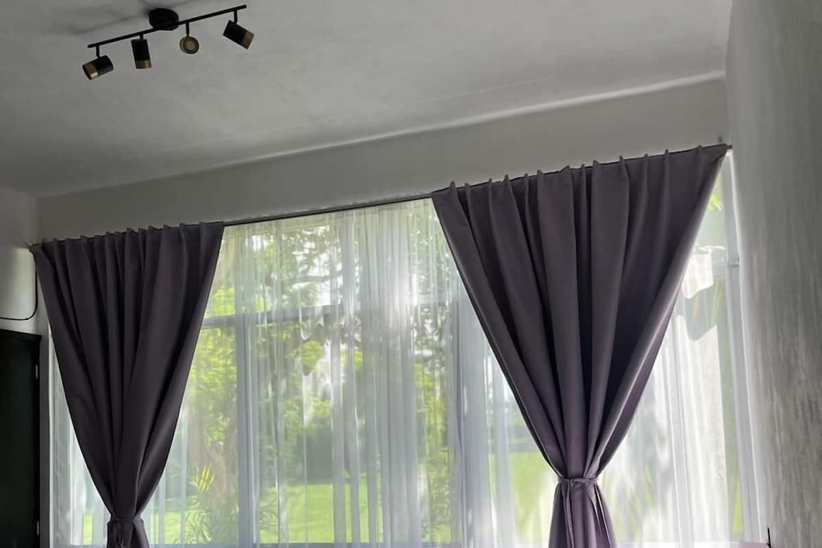 Ein Schlafzimmer mit einem Bett, zwei Fenstern mit violetten Vorhängen, einem Deckenventilator und Blick auf Grünfläche.
