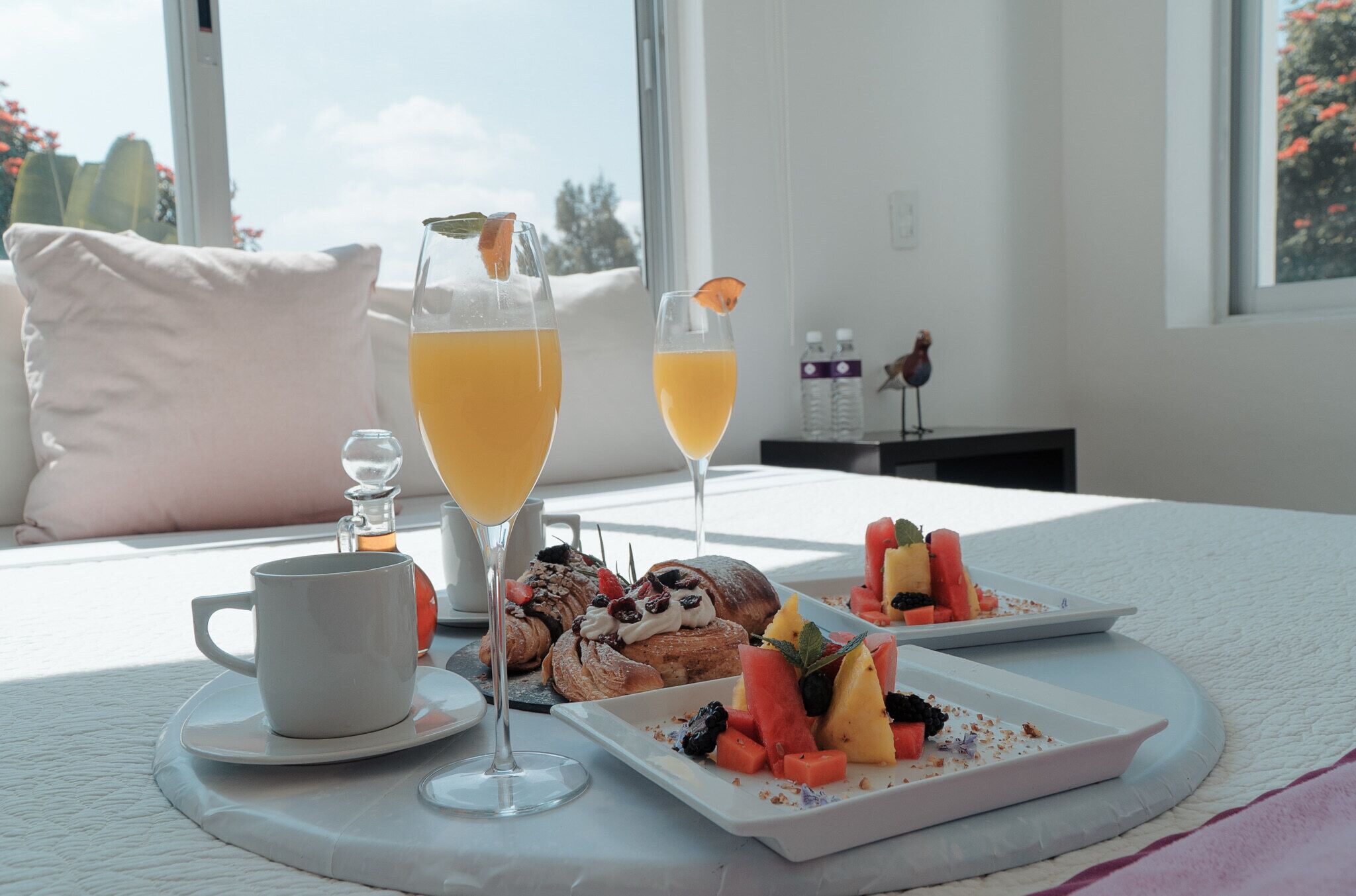 A breakfast setting with a white table, a cup, two glasses of orange juice, pastries, and fruit on plates.