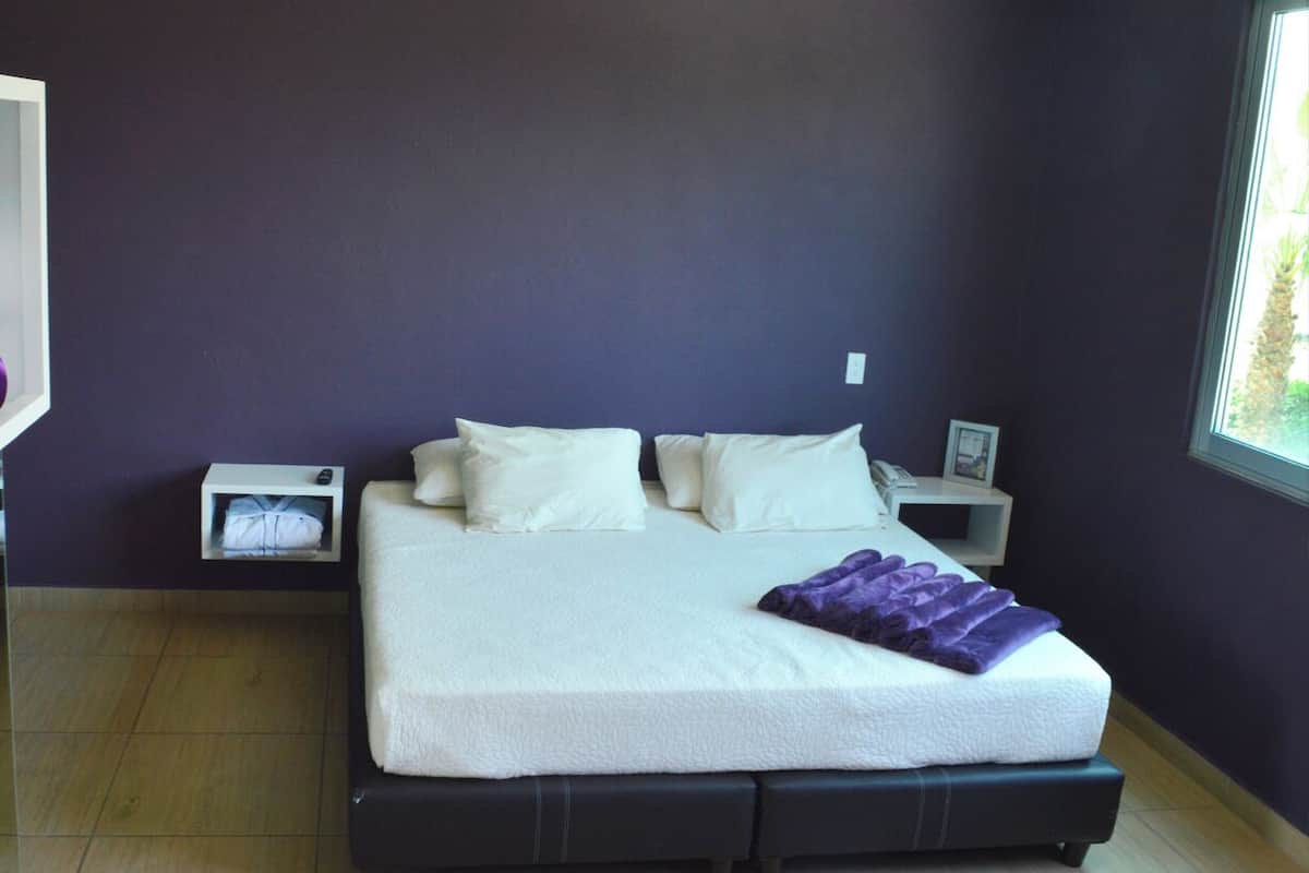Ein modernes Schlafzimmer mit einem großen Bett, zwei Nachttischen und einem Fenster mit Blick auf Palmen.