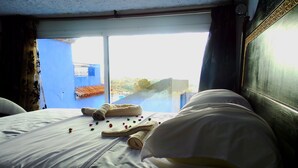 A hotel room with a bed, pillows, and a window with a view of buildings. - Riad Antek (Chefchaouen)